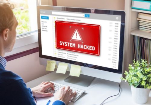 man sitting at a desktop computer wit system hacked on the screen